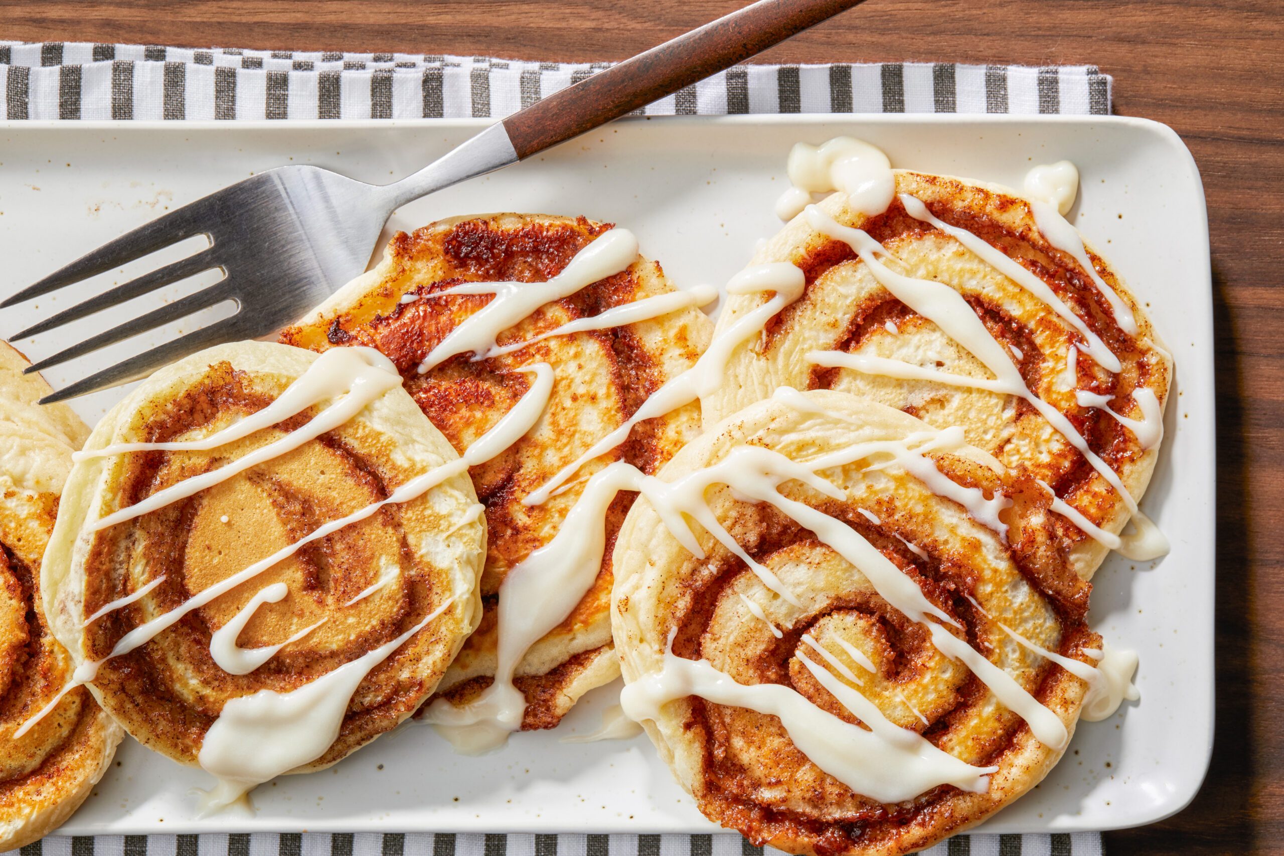 A plate of five cinnamon rolls drizzled with white icing, arranged neatly. A metal spatula rests on the plate, and the setup is on a wooden table with a striped cloth underneath.