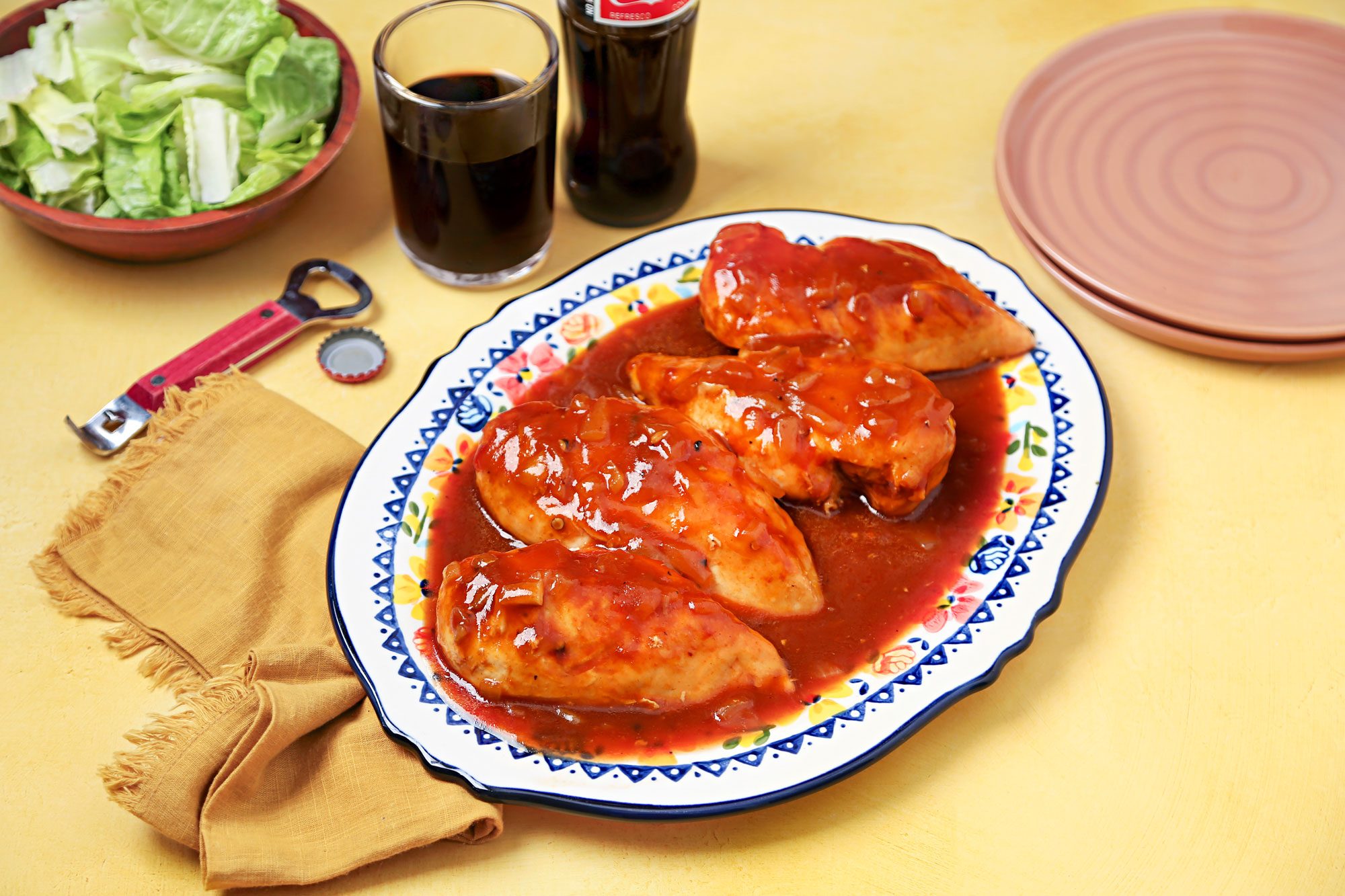 Shot of dish of cola chicken on a white plate with a floral design; the chicken is surrounded by a small amount of onion; there are two glasses of dark liquid, one glass of soda; and a bottle of soda on the table; there is also a bowl of salad and a bottle opener; the table is a light yellow color and there is a yellow napkin on the table