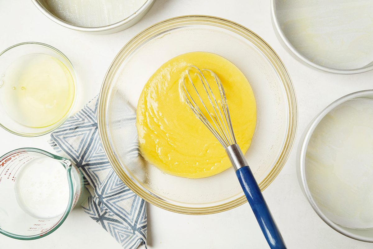 A glass bowl filled with yellow cake batter and a blue whisk on a white surface. Surrounding items include empty round baking pans, a textured gray cloth, and a measuring cup with a small amount of liquid.