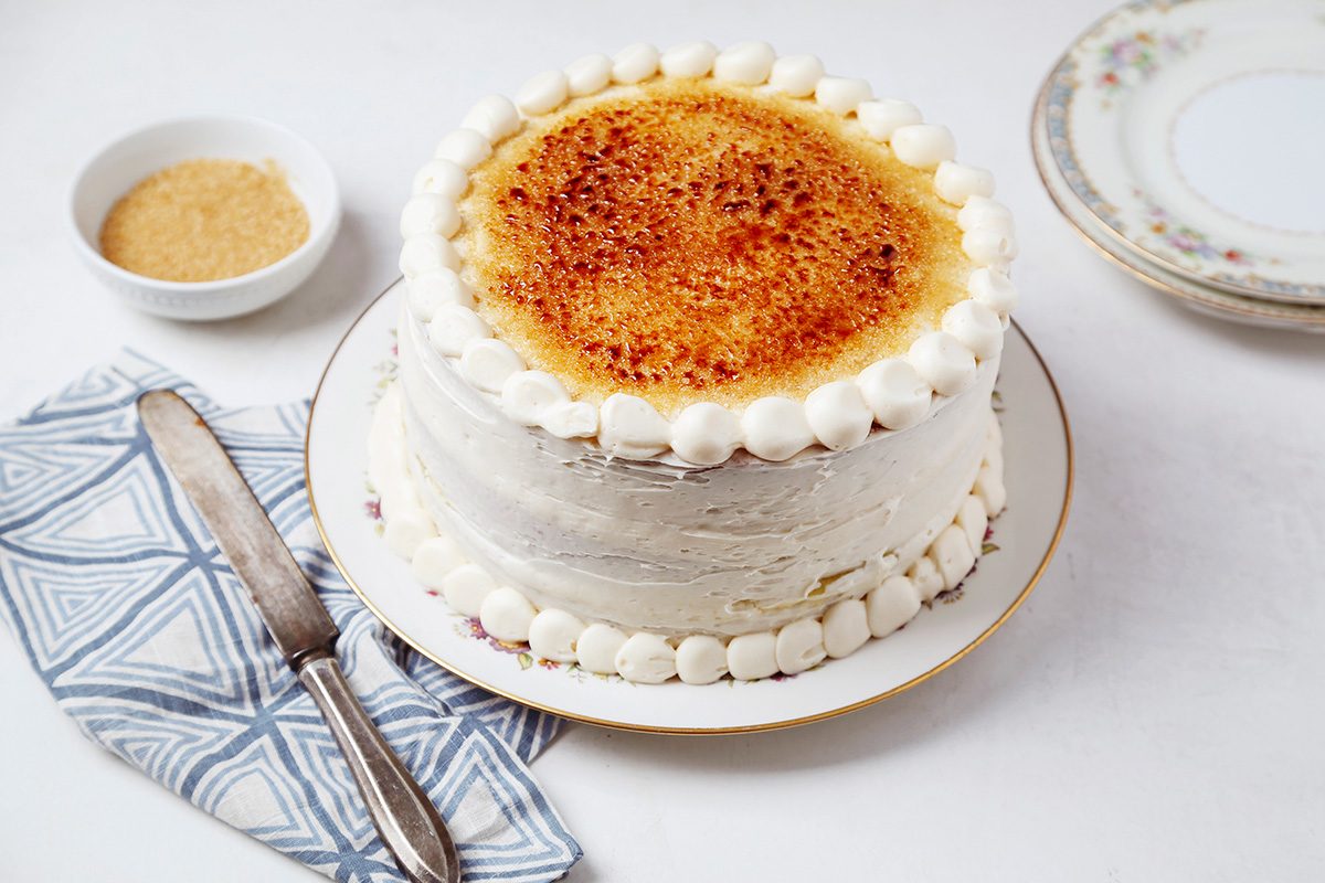 A layered cake with white frosting and a caramelized sugar top on a plate. It's surrounded by a napkin, a knife, and a bowl of brown sugar on a white table.