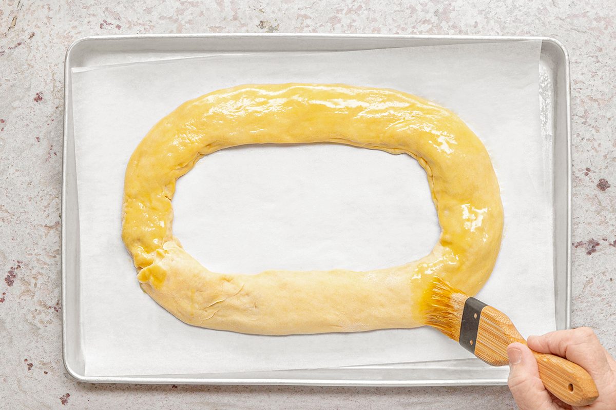 An oval-shaped dough ring on parchment paper is being brushed with a golden liquid using a wooden brush, on a baking tray.