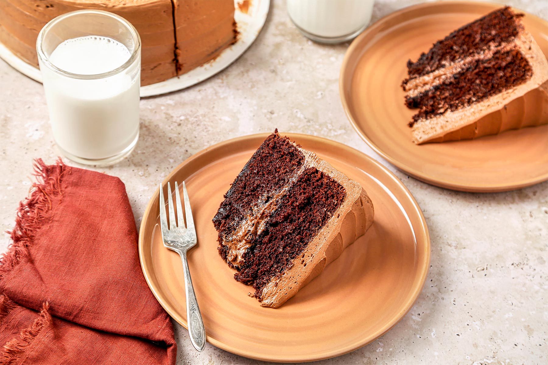 3/4th shot of Devil's Food Cake shot on a beige marble surface. Sliced cake served on tan plates with milk.