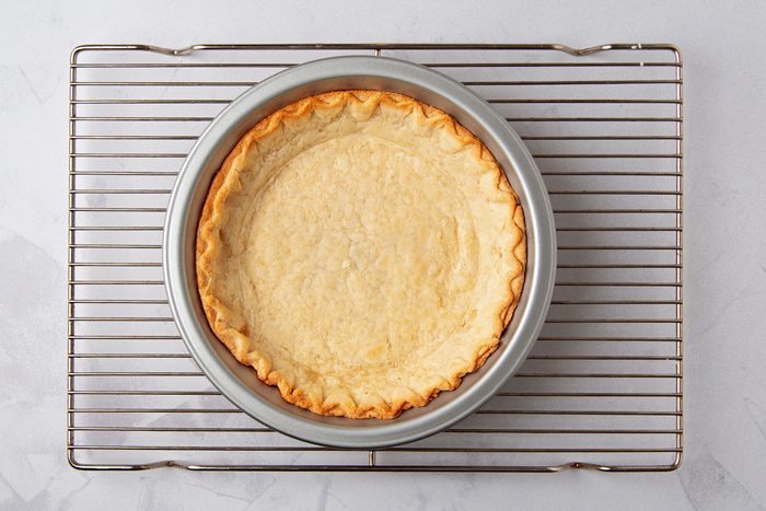 overhead shot of a pie crust in a pie pan on a silver cooling rack, against a white background; the pie crust is golden brown and has a crimped edge; the pie pan is a light silver color