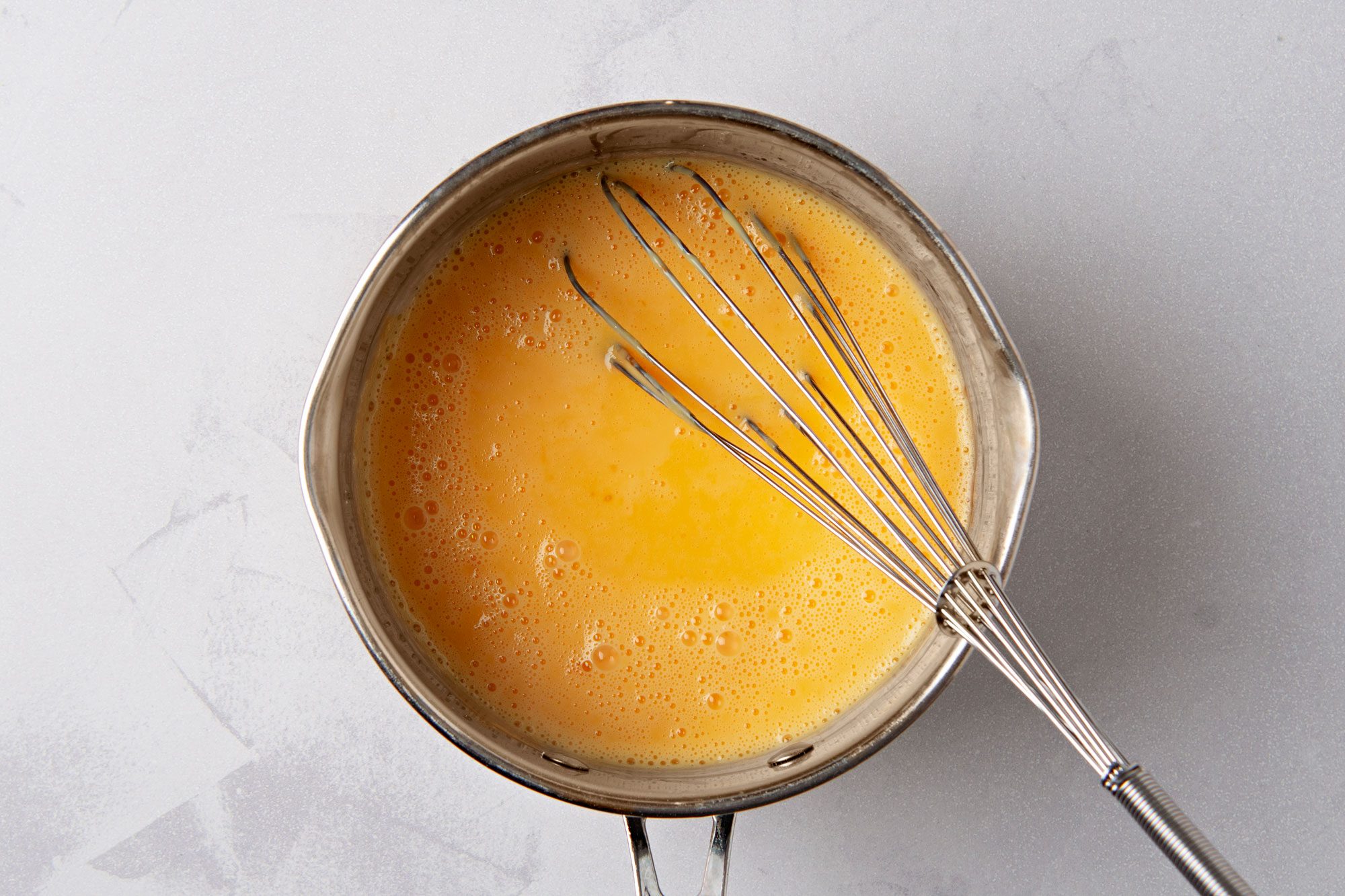 overhead shot of a stainless steel saucepan with a whisk resting in it, filled with a light orange liquid; the liquid has tiny air bubbles on the surface; the saucepan is on a white surface