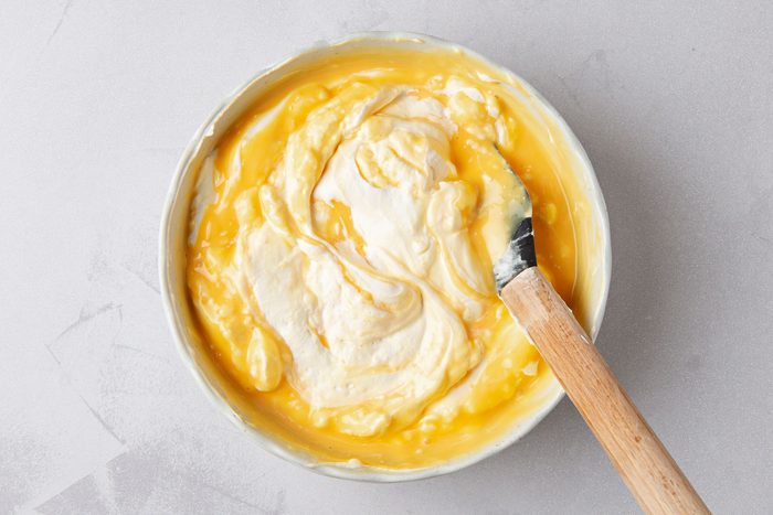 overhead shot of a white bowl filled with a creamy yellow mixture, a wooden spatula rests on the edge of the bowl, partially submerged in the mixture; the bowl sits on a grey textured surface