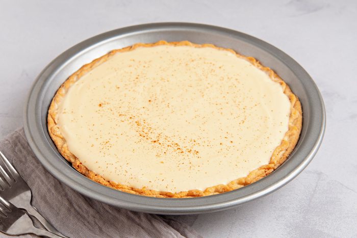 3/4th shot of a pie in a silver pie pan; the pie is filled with a light yellow custard-like filling, and it is sprinkled with a brown spice; the crust is golden brown; the pie is sitting on a white surface and there are two silver forks next to it on a grey napkin