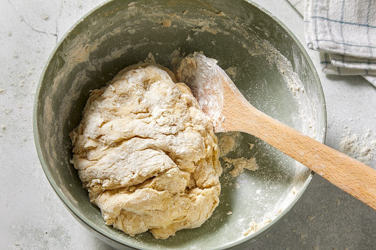 A mixing bowl containing a partially kneaded dough with a wooden spoon resting on the side. 