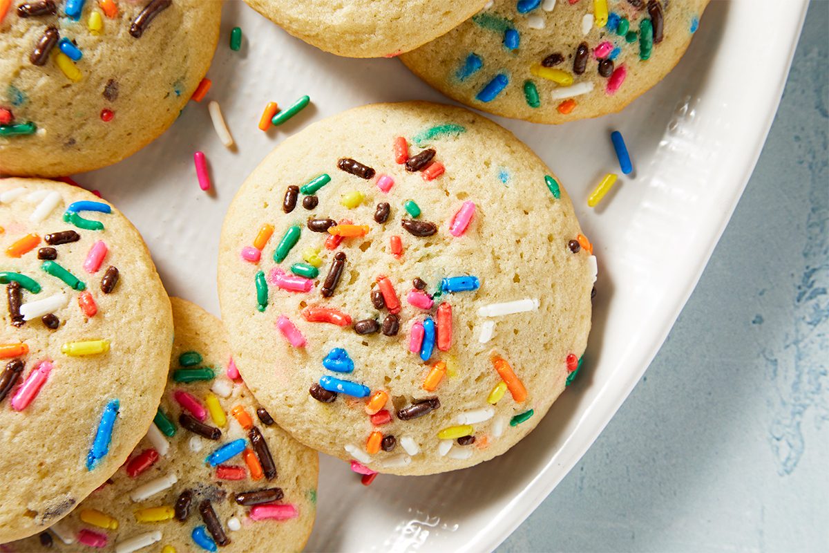 Close-up of colorful sprinkle-topped cookies on a white plate. The cookies are light brown and decorated with multicolored sprinkles, including blue, pink, yellow, and brown, creating a festive look. The background is a light gray surface.