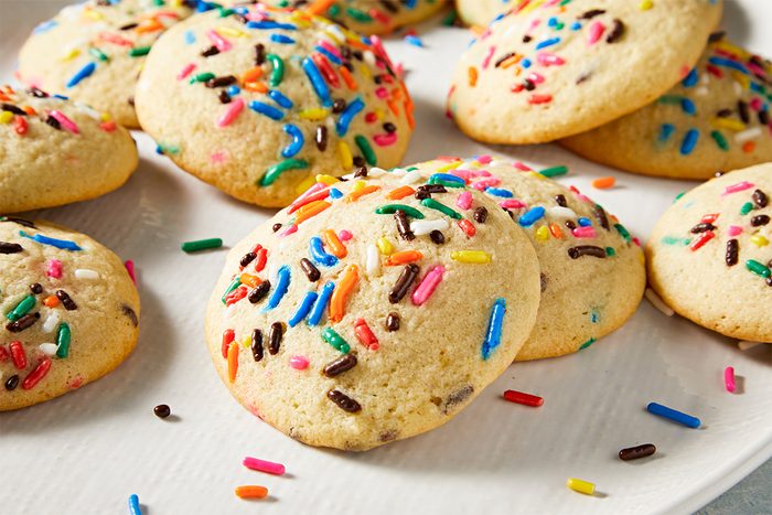 A plate of round, soft cookies topped with colorful sprinkles. The cookies are light golden brown, scattered with red, green, yellow, blue, and chocolate sprinkles, set against a simple, light-colored background.