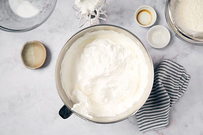 A mixing bowl filled with whipped cream on a countertop. Surrounding it are ingredients in small bowls, including sugar and vanilla extract. A striped cloth lies nearby, and a sieve with flour is partially visible.