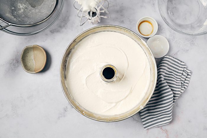 A tube pan filled with cake batter on a countertop, surrounded by a striped cloth, a fine-mesh strainer, and small bowls containing different ingredients.