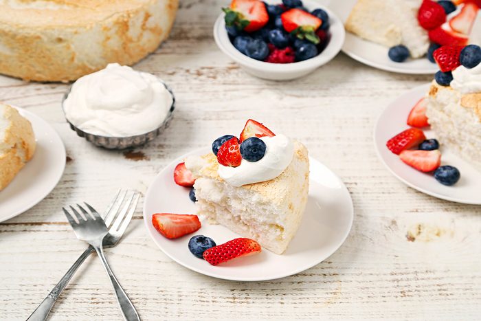 Slice of angel food cake topped with whipped cream and berries on a white plate. Fresh strawberries and blueberries are scattered around. Additional plates with cake and bowls of berries are on a rustic white wooden table. Two forks are placed nearby.