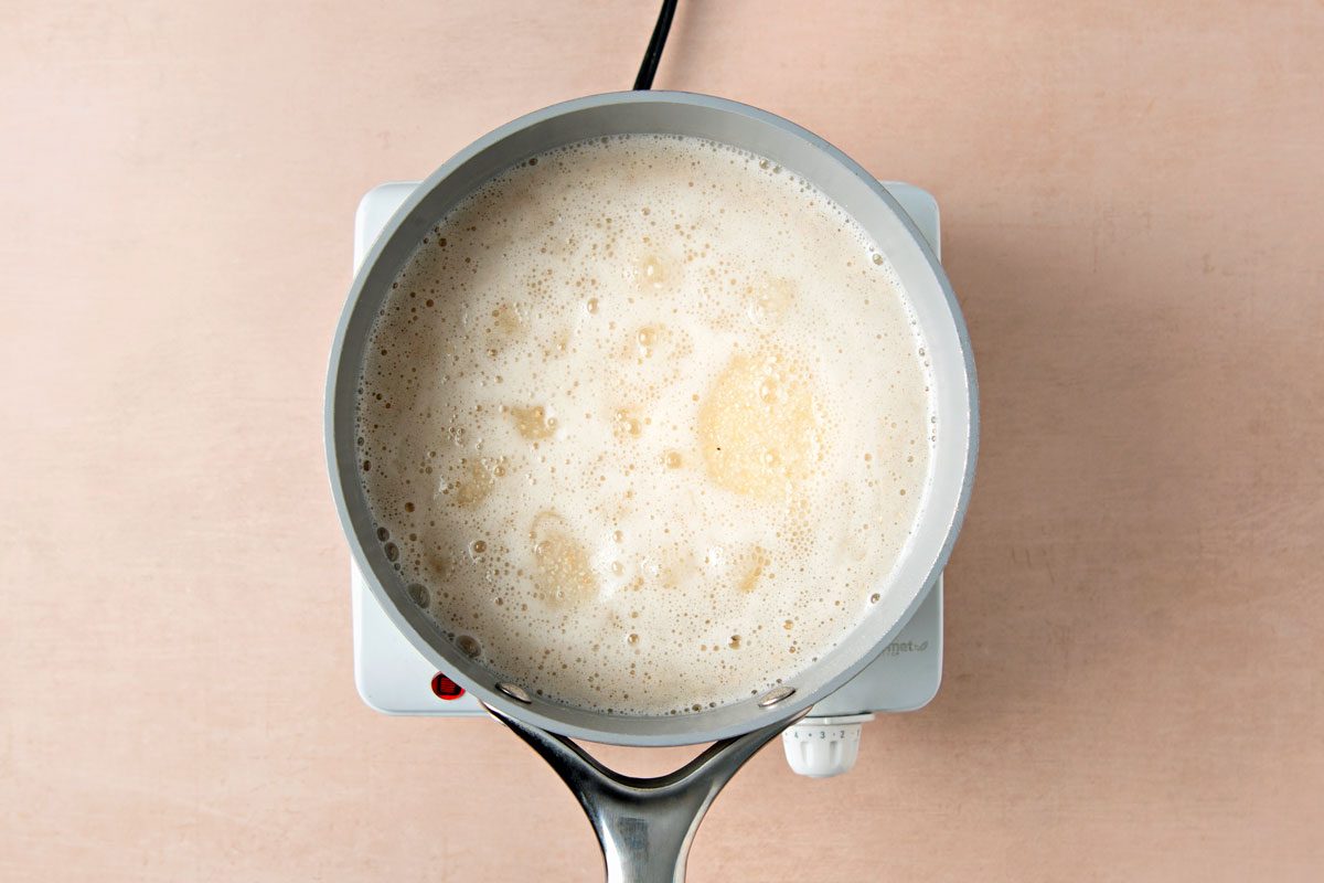 overhead shot of a saucepan with a silver handle on a white burner; the saucepan is filled with a white, foamy liquid; the burner is on a light brown countertop