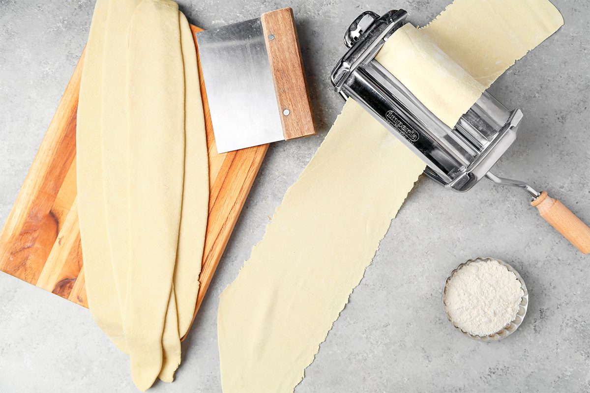 A pasta maker with a wooden handle rolls out sheets of dough on a gray countertop. A wooden cutting board with dough strips and a bench scraper are nearby. A small bowl of flour sits beside the pasta maker.