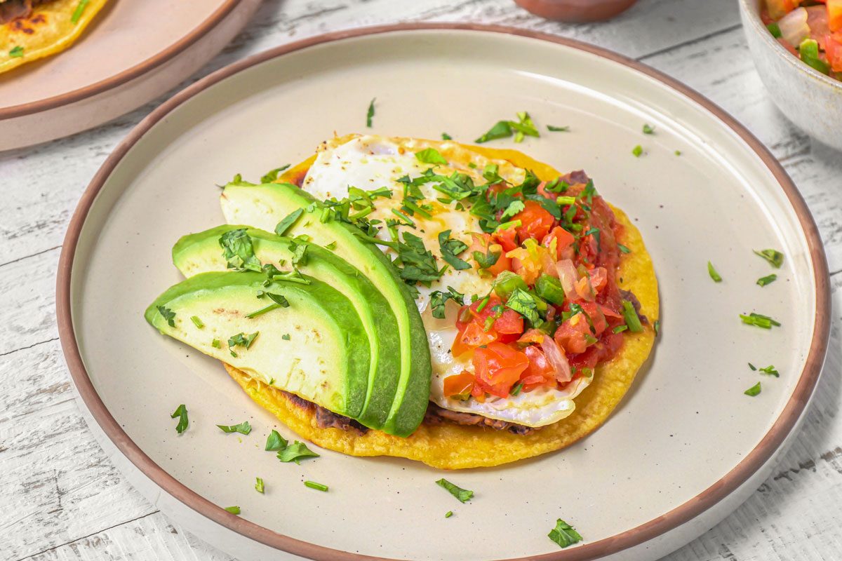 3/4th shot of Huevos Rancheros; it consists of a fried egg, a corn tortilla, salsa, avocado and cilantro; the tortilla is topped with refried beans, a fried egg, and a generous amount of salsa; the salsa is made of diced tomatoes, onions, and cilantro; avocado slices are fanned out around the egg and tortilla