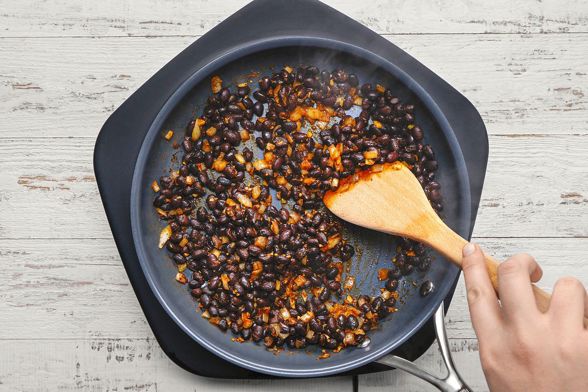 overhead shot of a person is cooking black beans and onions in a frying pan; the pan is sitting on a white wooden surface