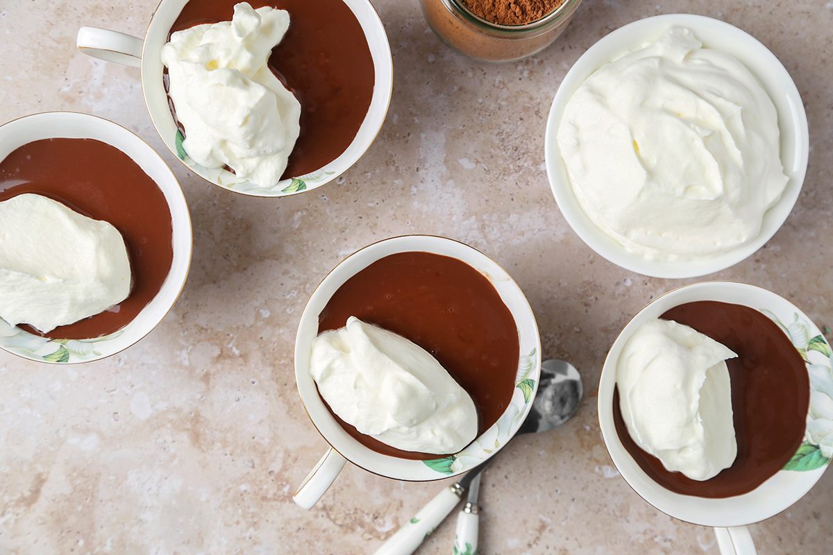 Top view of four cups of chocolate pudding topped with whipped cream, arranged on a light marble surface. A bowl filled with more whipped cream and a jar of toppings are nearby, with two spoons beside the cups.
