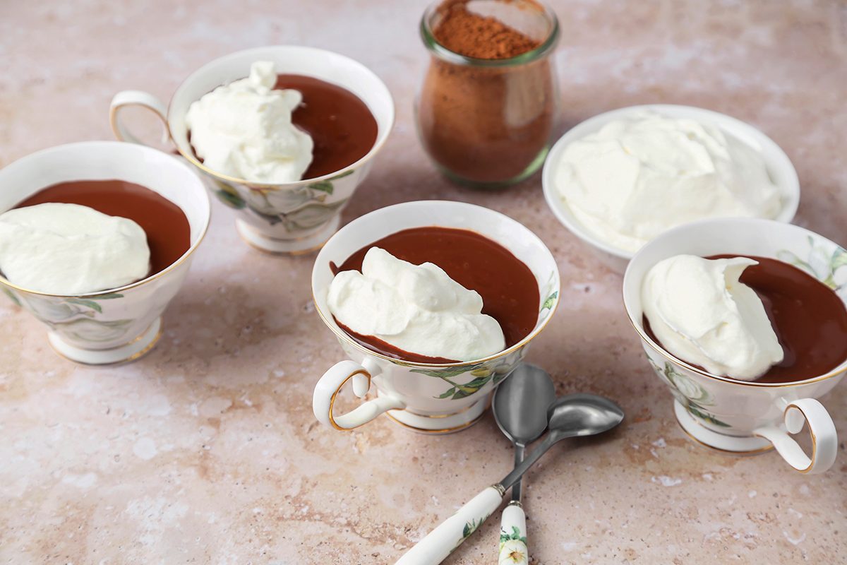 Four cups of chocolate pudding topped with whipped cream are arranged on a table. Two spoons lie in front of the cups. A jar with cocoa powder and a small plate with whipped cream are in the background.