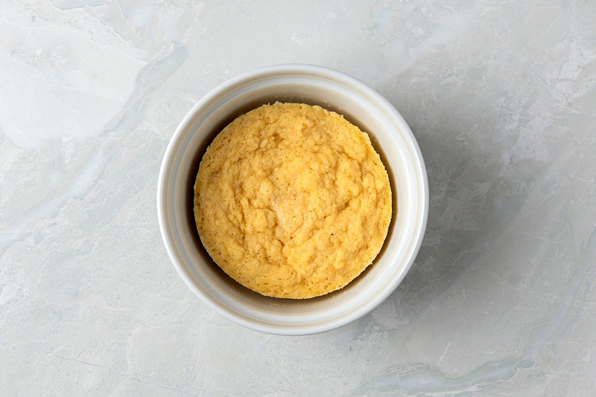 A round, fluffy cornbread muffin sits in a white ramekin on a light gray marble background.