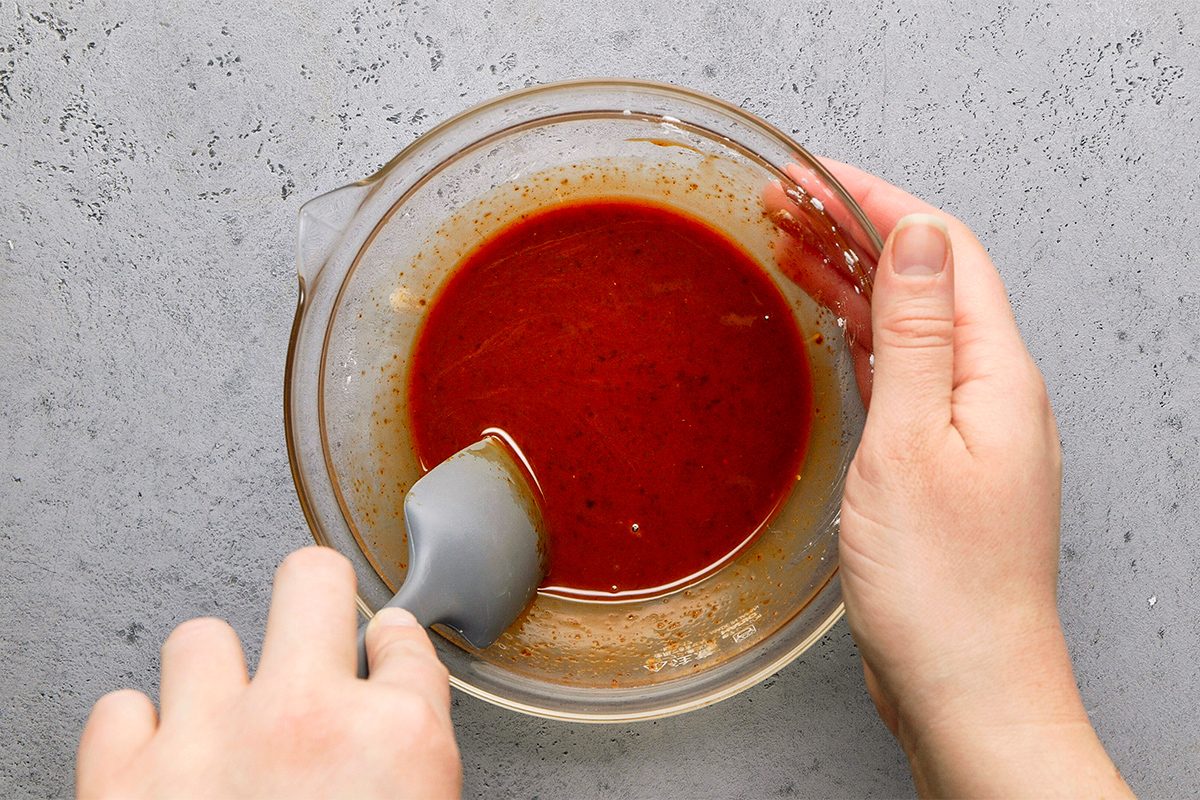 Hands holding a glass bowl filled with a red sauce, using a spatula to stir. The background is a gray, textured surface.