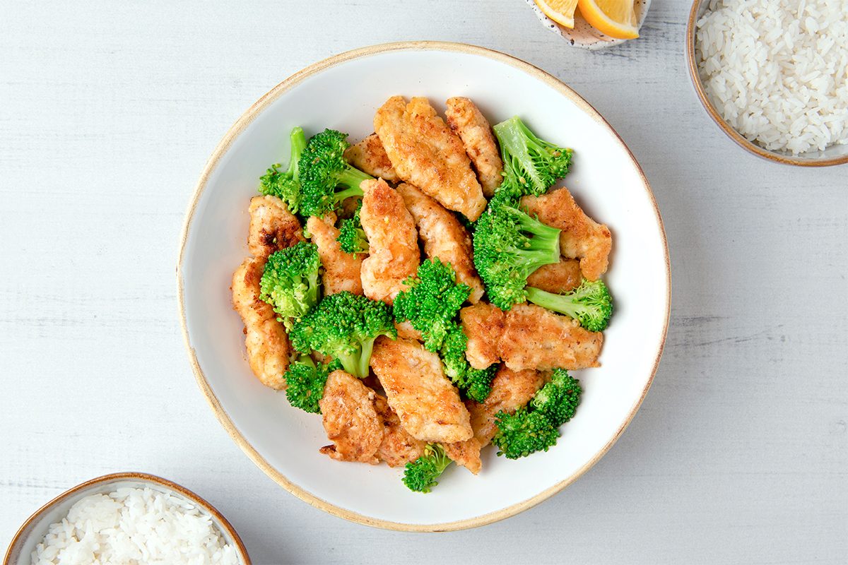 A bowl of fried chicken pieces mixed with broccoli florets. There are side bowls containing white rice and lemon wedges on a white wooden surface.