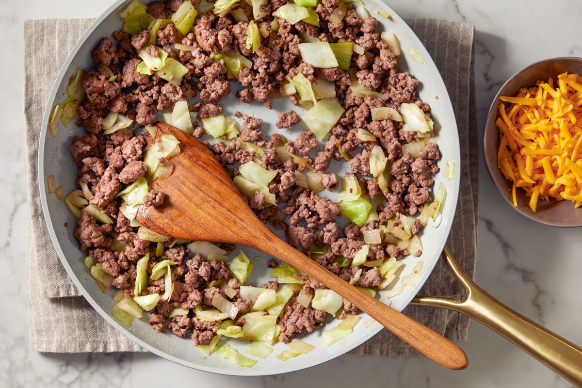 overhead shot of a frying pan filled with a mixture of cooked ground beef and chopped green cabbage; a wooden spatula is resting inside the pan, alongside the pan, there is a small bowl containing shredded cheddar cheese; the blurred background reveals a light marble surface, while the pan sits on a striped cloth