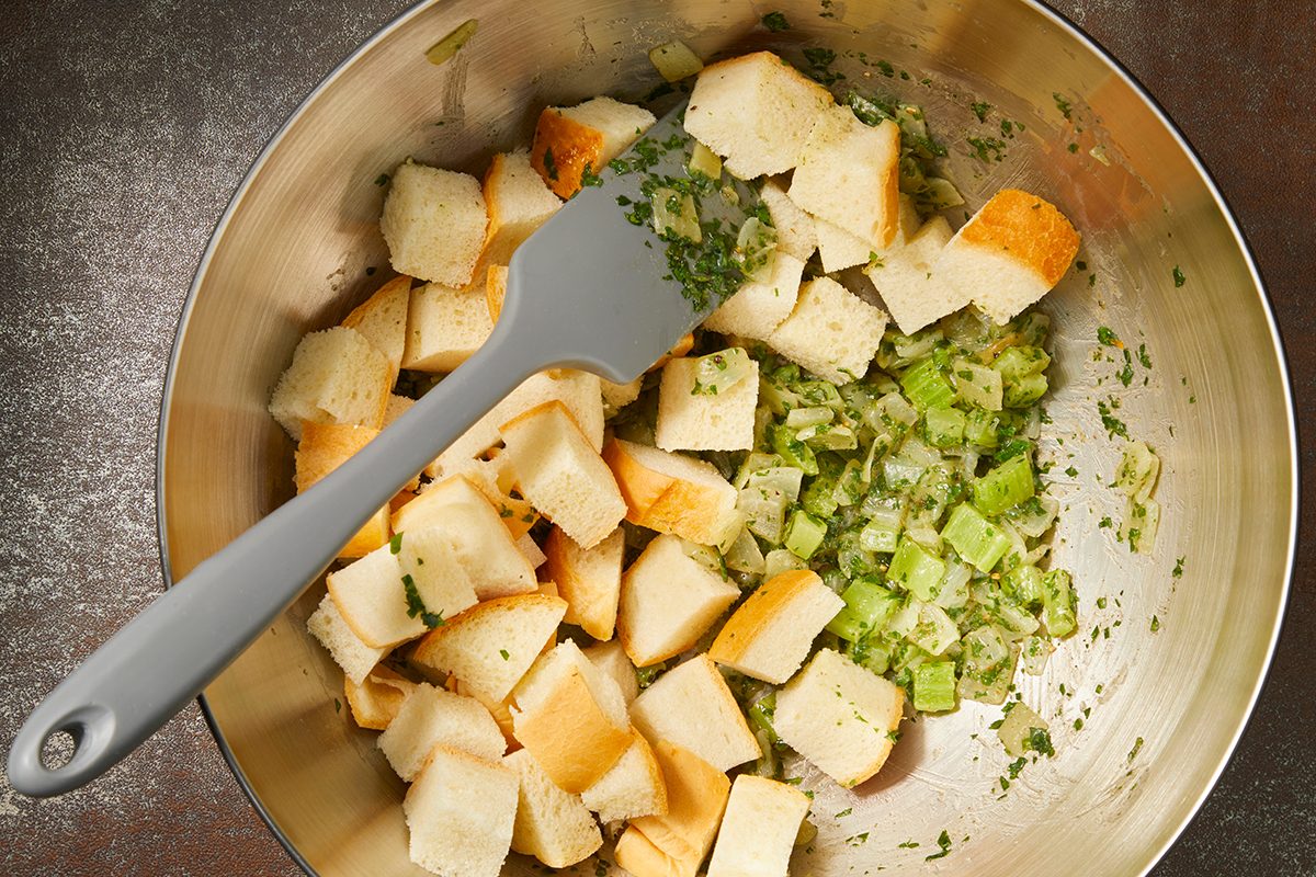 A stainless steel bowl containing cubed bread and chopped celery mixed with herbs. A gray spatula rests inside, partially covered by the ingredients. The bowl sits on a metallic surface.