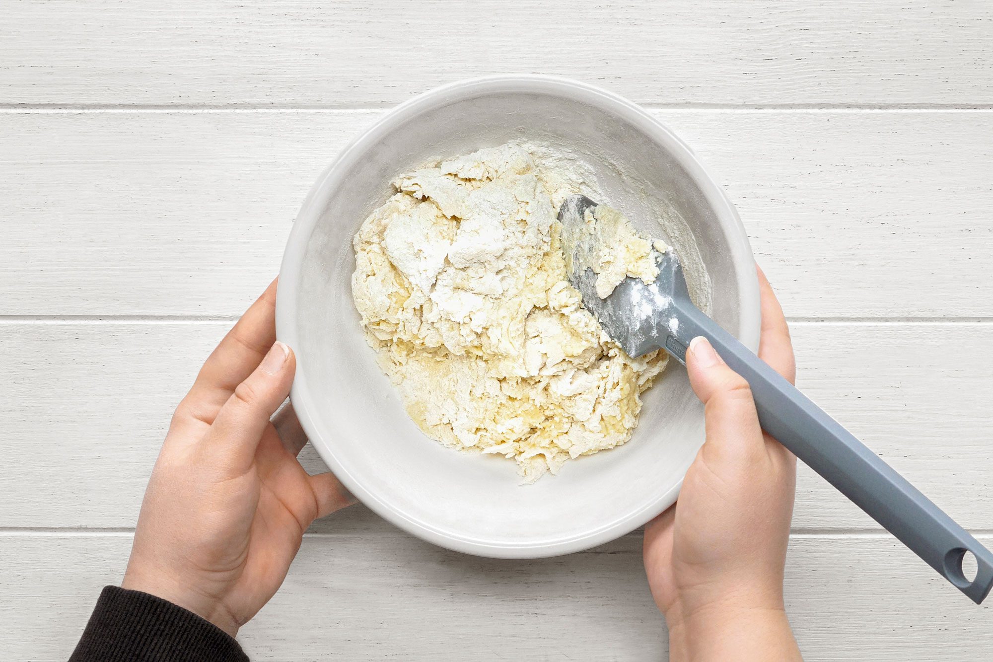 overhead shot of a pair of hands are holding a white bowl containing a mixture of dough and flour; a grey spatula is on top of the dough; the dough is being mixed; the bowl is being held by one hand; the spatula is being held by the other hand; the hands are on a white wooden background