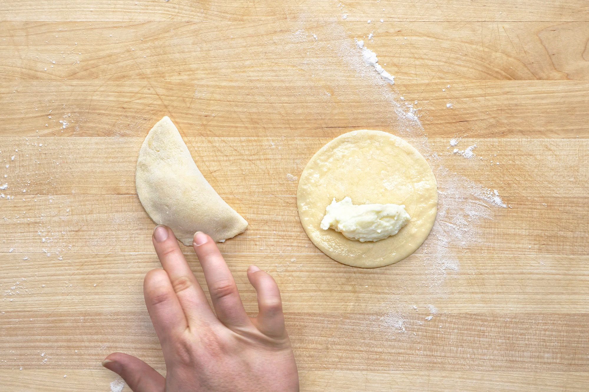 overhead shot of a wooden cutting board with two pieces of dough on it, a hand is seen in the lower left corner of the image, a half moon shaped dough is lying on the board and a round piece of dough is lying next to it, on top of the round piece of dough there is a filling which is white in colour; on the left side of the image there is a small amount of flour scattered on the board