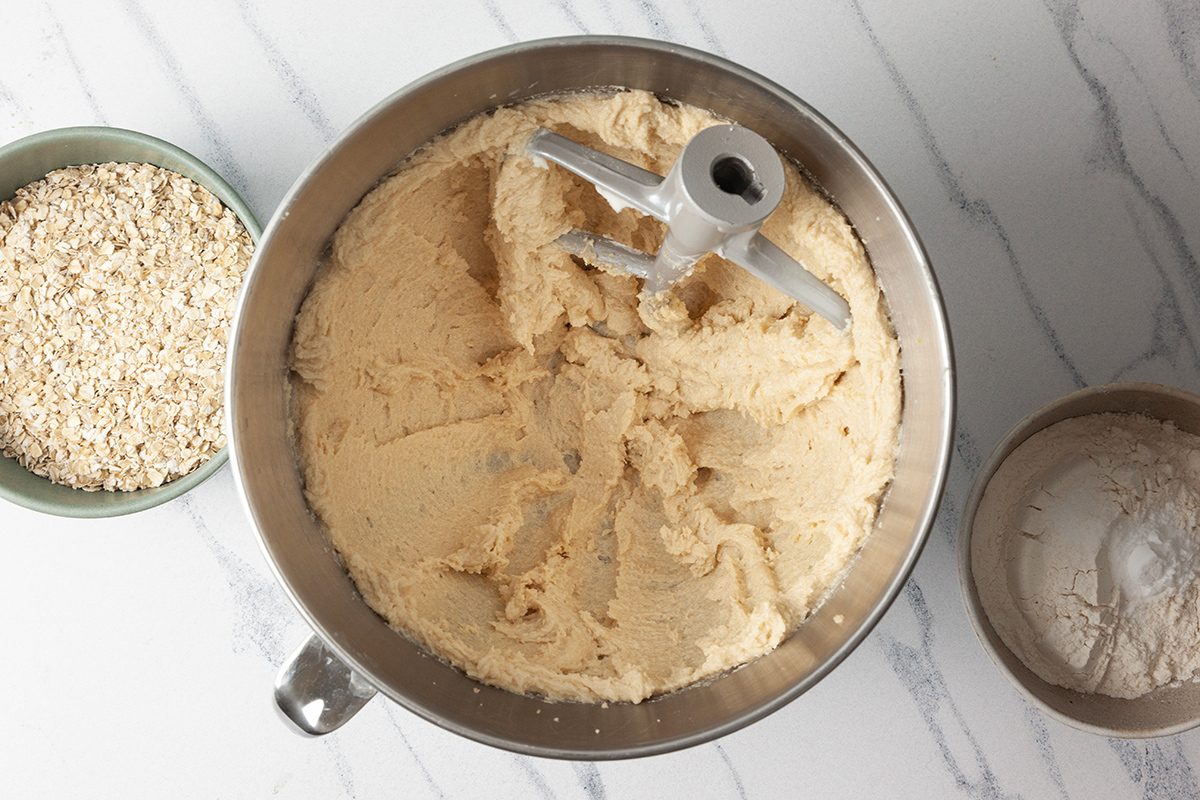 A mixing bowl filled with creamy dough, equipped with a paddle attachment, sits on a marble countertop. To the left, a bowl of oats is visible, and to the right, a bowl of flour.