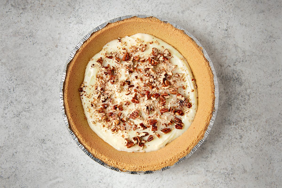 A pie with a graham cracker crust, filled with creamy white filling, topped with crumbled pieces of pecans, sits on a textured gray surface.