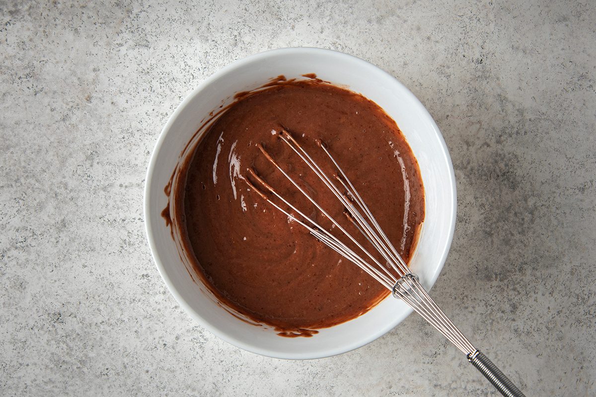 A white bowl filled with chocolate cake batter on a gray textured surface. A metal whisk rests inside the bowl.