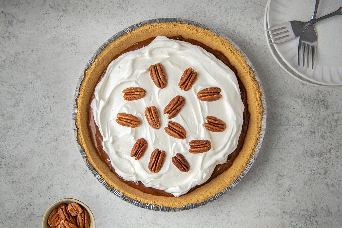 A pie with a graham cracker crust, topped with whipped cream and arranged pecans. A small bowl with pecans is on the side, and there is a stack of plates with forks in the background.