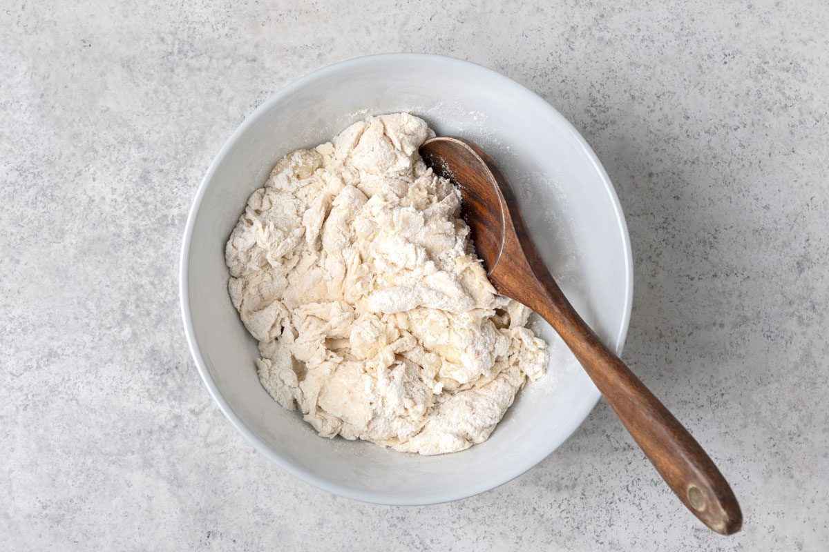 overhead shot of a light grey bowl filled with a lumpy, white mixture that appears to be dough, a wooden spoon rests inside the bowl, partially submerged in the dough, the background is a mottled grey surface;