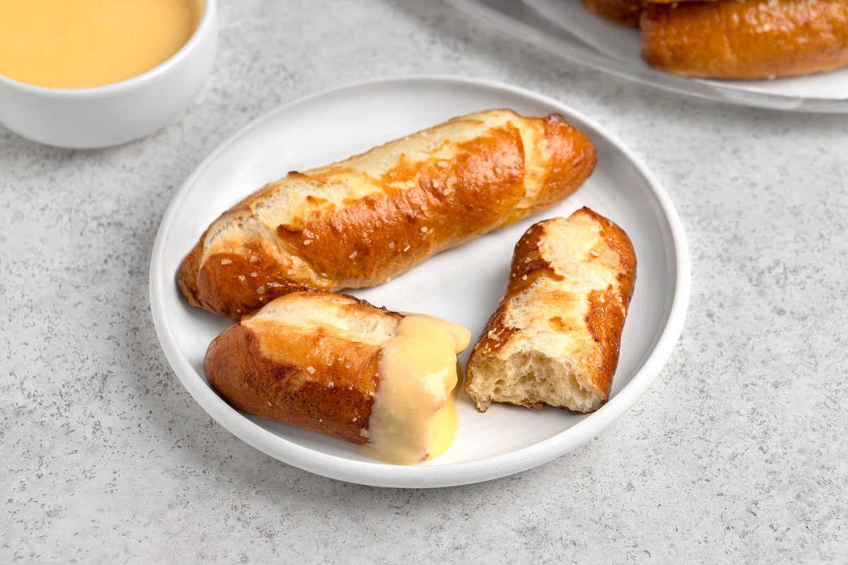 3/4th shot of three pretzel rolls on a white plate, two of the rolls are whole and one is partially eaten revealing the inside texture, one of the whole rolls is drizzled with a yellow cheese sauce, in the upper left corner, a white bowl filled with the same yellow cheese sauce is partially visible, the background is a light gray countertop or surface;