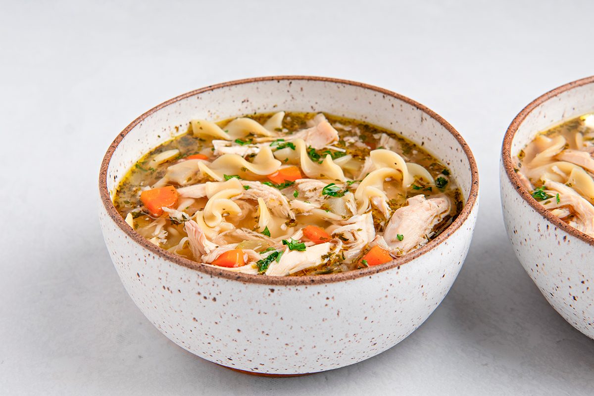 A bowl of chicken noodle soup containing shredded chicken, egg noodles, diced carrots, and chopped parsley. The soup is served in a speckled white bowl on a light-colored surface.