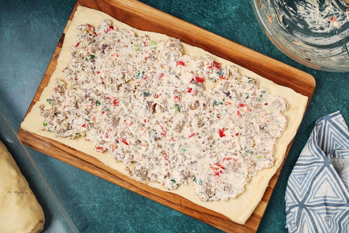 overhead shot of a wooden cutting board with a sheet of dough on it, topped with a mixture of cream cheese, sausage, red bell peppers, and green onions; the cutting board is on a teal surface;