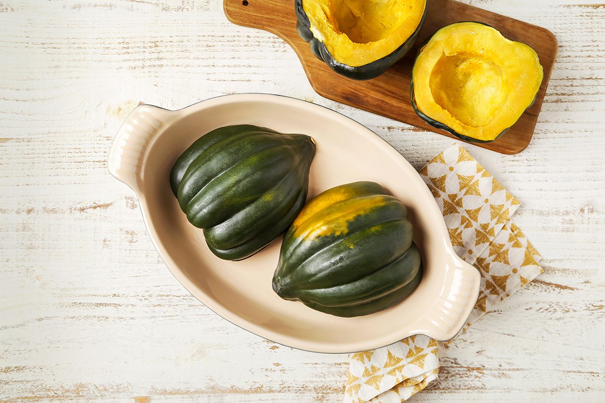 Two whole acorn squashes sit in a cream-colored baking dish on a wooden table. Nearby, two squash halves rest on a wooden board, revealing their orange interiors. A patterned napkin is placed beside the dish.