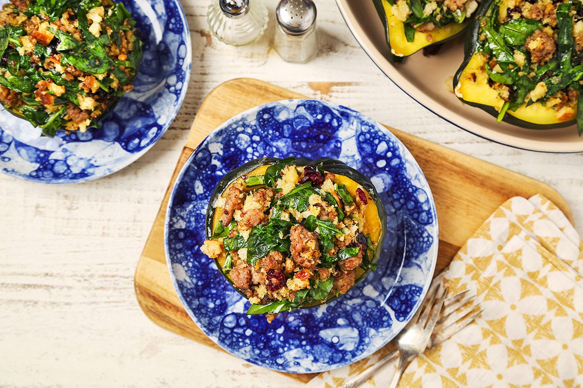 A blue and white plate holds a stuffed acorn squash filled with a mixture of spinach, quinoa, cranberries, and crumbled cheese. It's served on a wooden board with cutlery and a napkin nearby. Other plates with similar dishes are partially visible.
