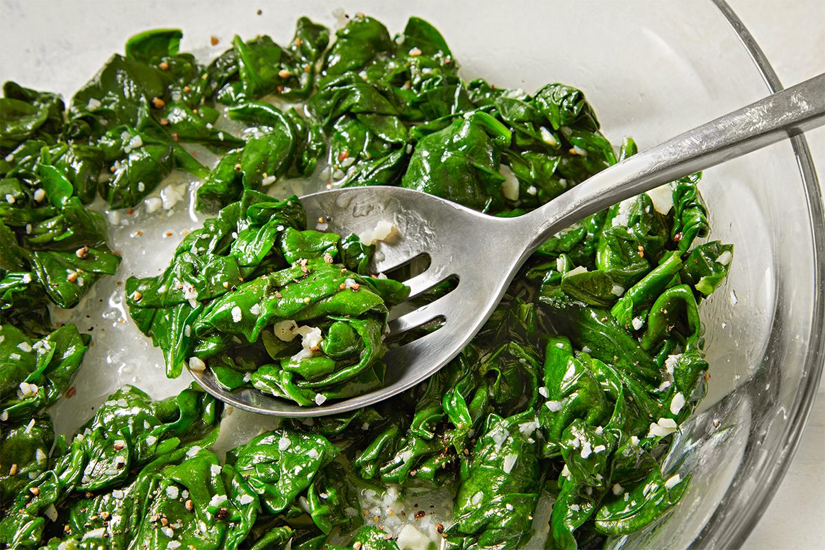 A close-up of a bowl containing fresh spinach leaves coated with sesame seeds and a light dressing. A metal spoon is resting on the spinach, suggesting the dish is ready to be served.