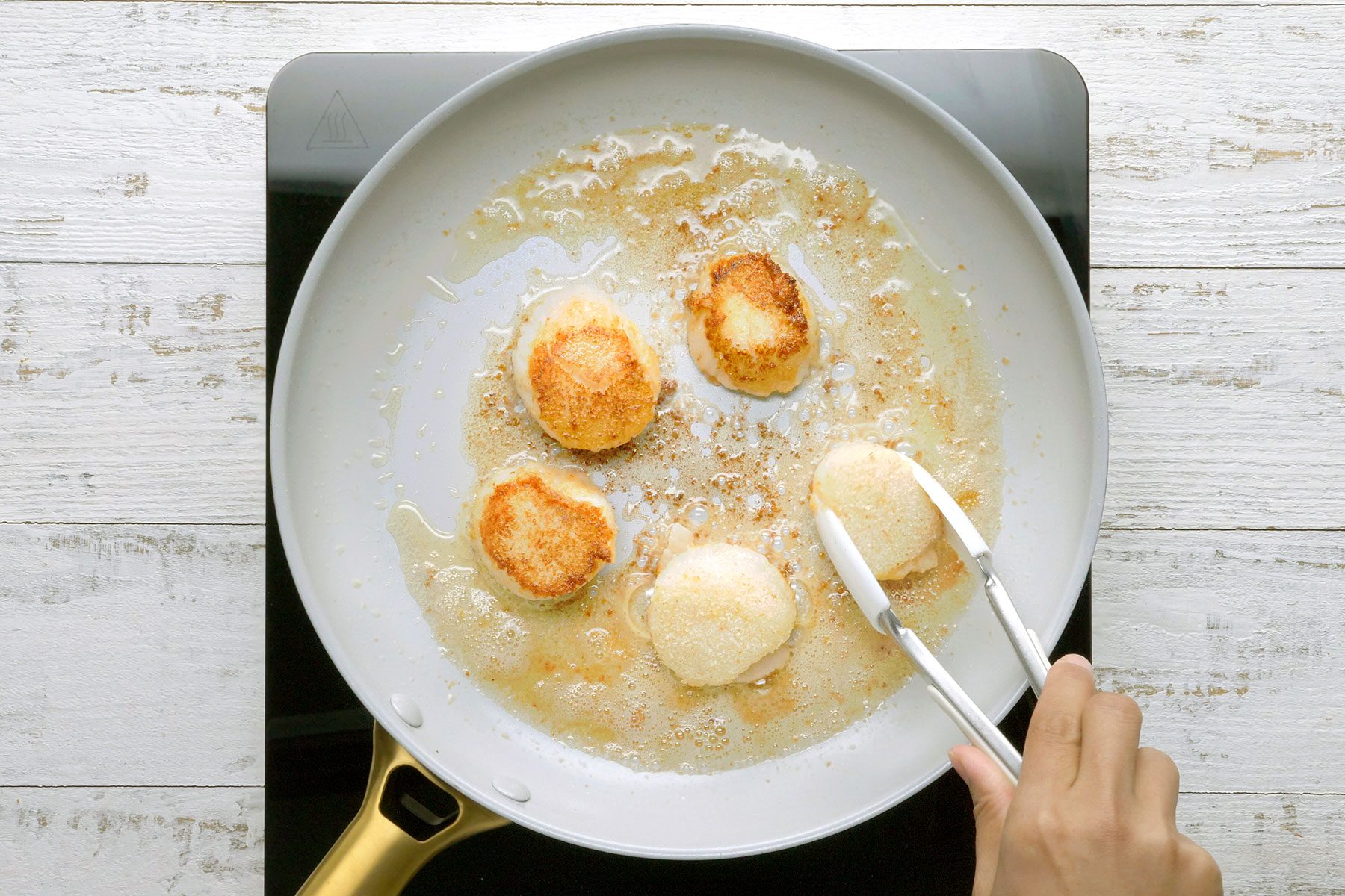 A person uses tongs to flip scallops in a frying pan on a stovetop. The scallops are golden brown and sizzling in oil. 
