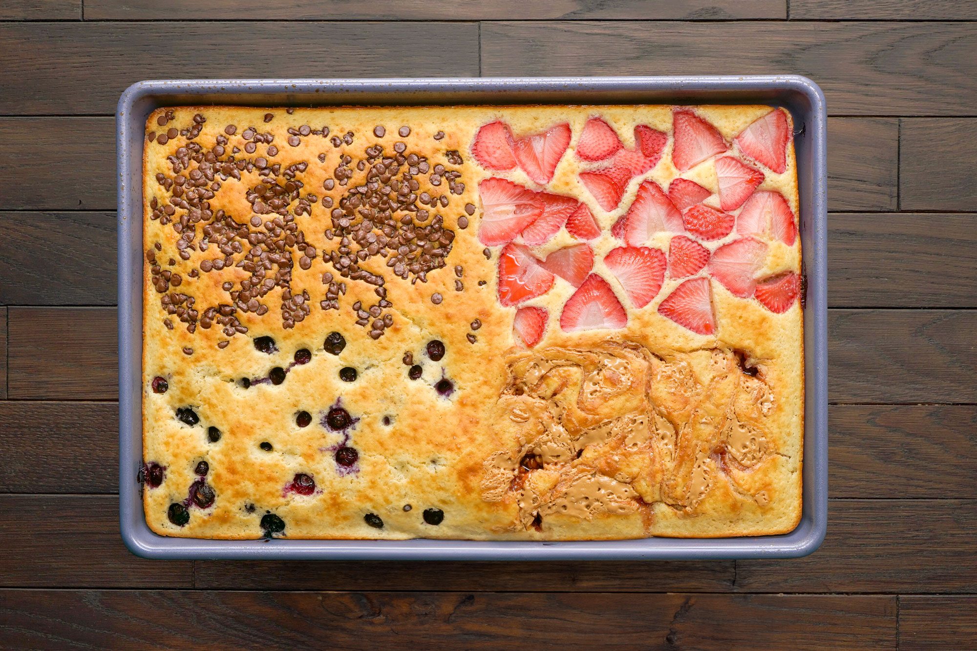 overhead shot of a large rectangular sheet pan filled with pancakes; variety of toppings; one section is adorned with chocolate chips, while another features bright red strawberry slices, there are also blueberries scattered across, wooden plank background