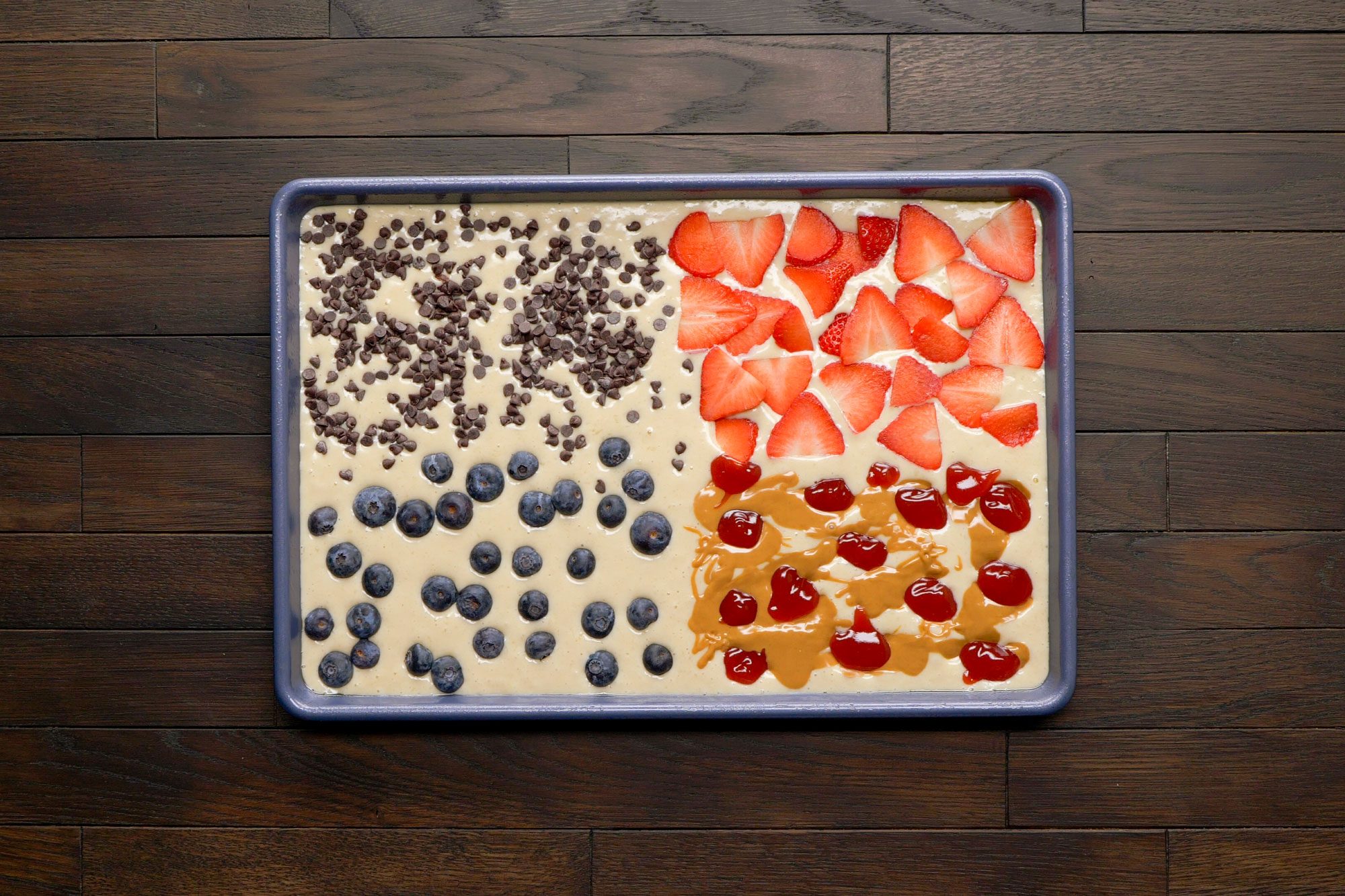 overhead shot of a baking tray filled with a pancake mixture that is divided into sections; one section is topped with chocolate chips; another features fresh blueberries; a third section is adorned with sliced strawberries; the final area includes streaks of peanut butter and a drizzle of syrup; over dark wooden surface