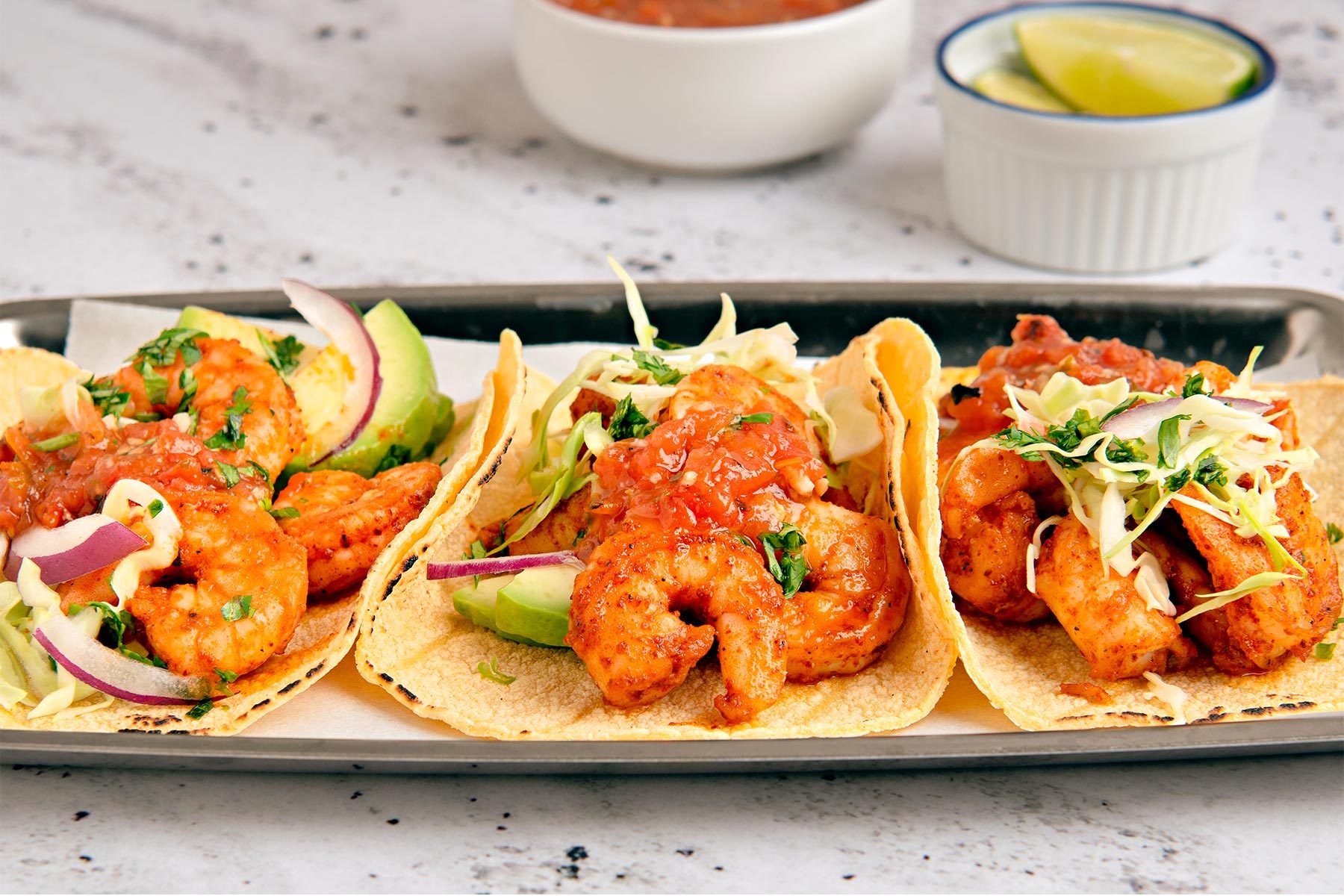 3/4th shot of three shrimp tacos on a white surface, two bowls are slightly out of focus in the background, one containing red salsa and the other holding lime wedges ;