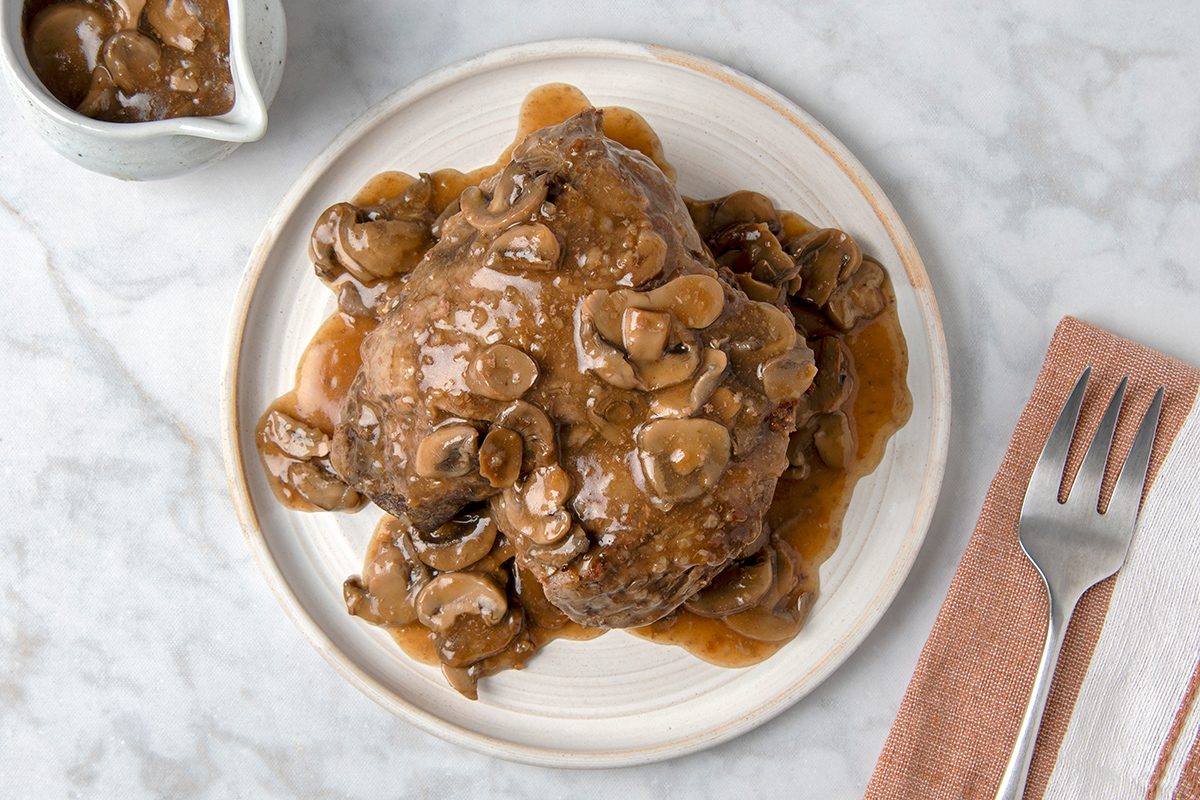 A plate of Salisbury steak covered in mushroom gravy, sitting on a marble surface. A napkin with a fork and knife is placed beside the plate.