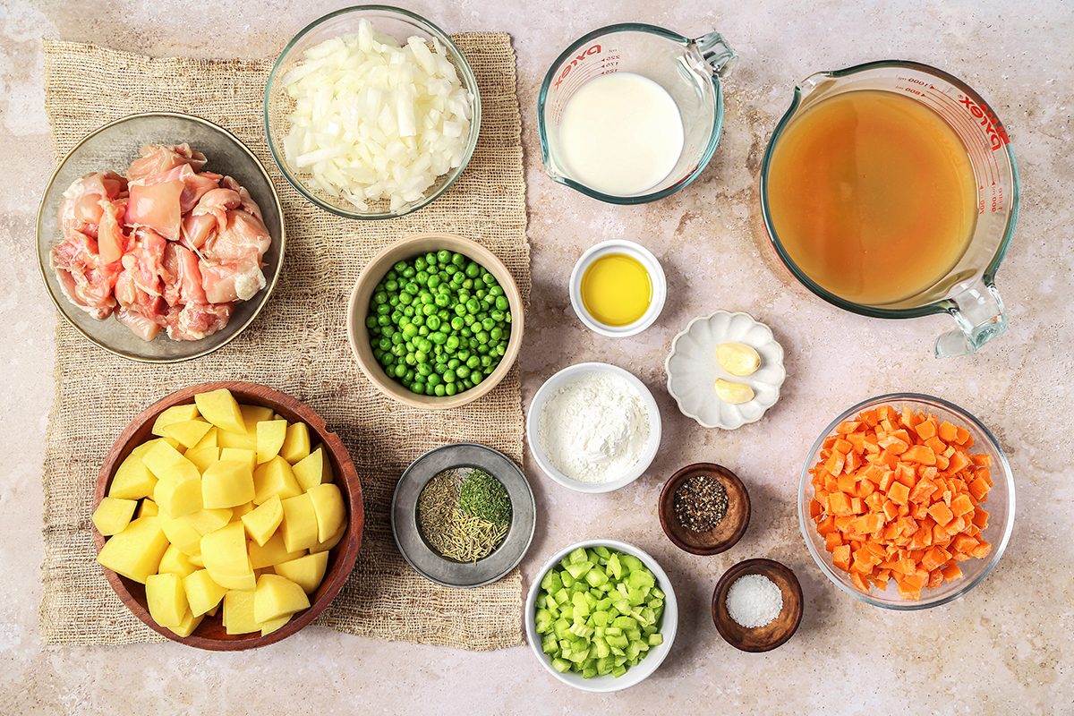 Ingredients arranged on a countertop include cubed potatoes, diced chicken, onions, peas, carrots, celery, garlic, flour, herbs, pepper, oil, cream, and broth.