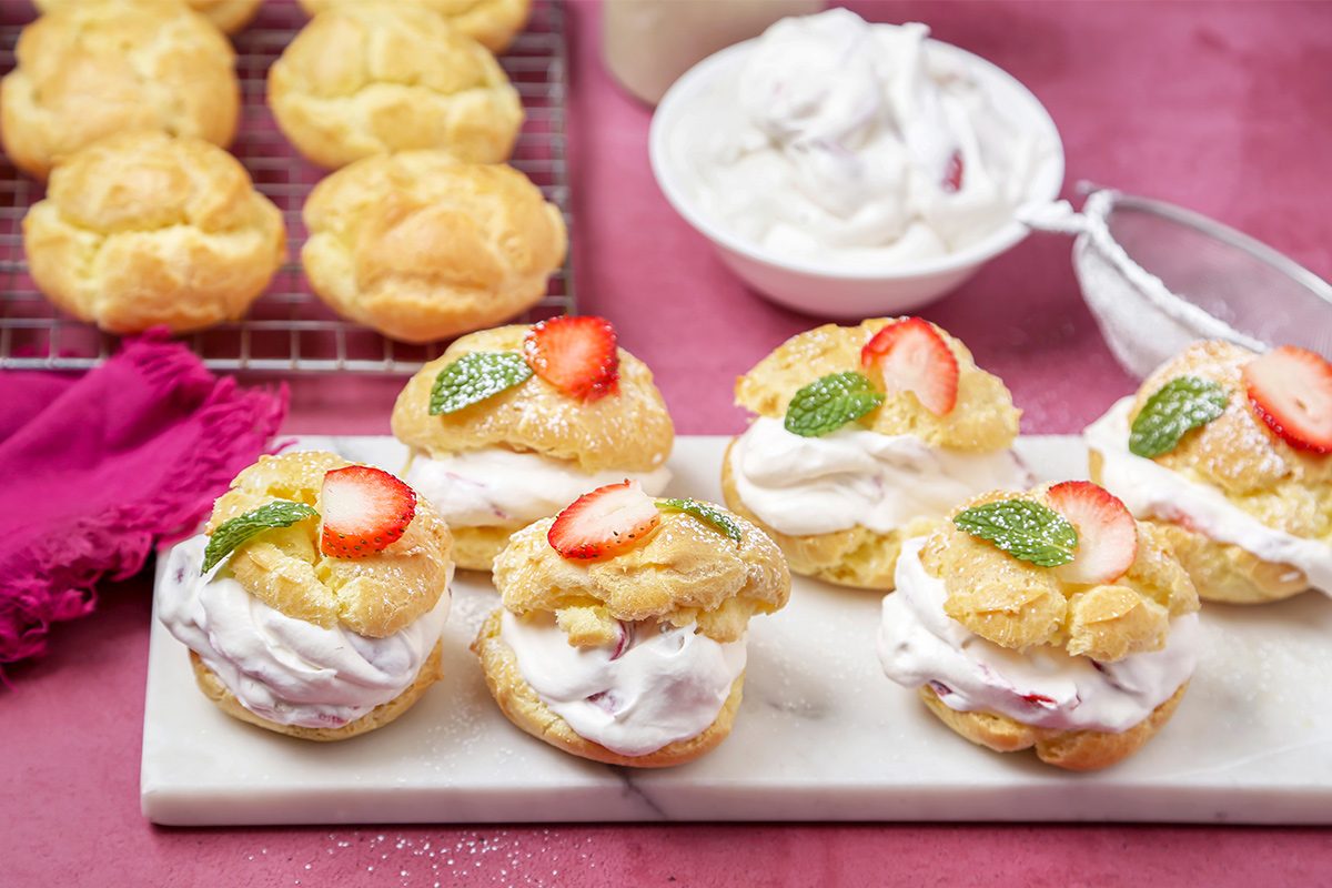 Side view of garnished strawberry cream puffs served next to the puffs kept on a cooling tray, cream, and a pink napkin on a pink tabletop.