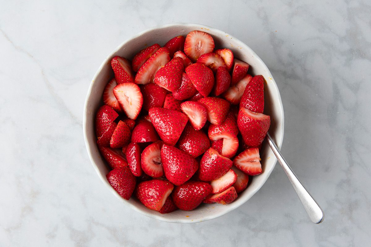 A bowl filled with fresh, sliced strawberries on a light marble surface. A spoon rests inside the bowl.