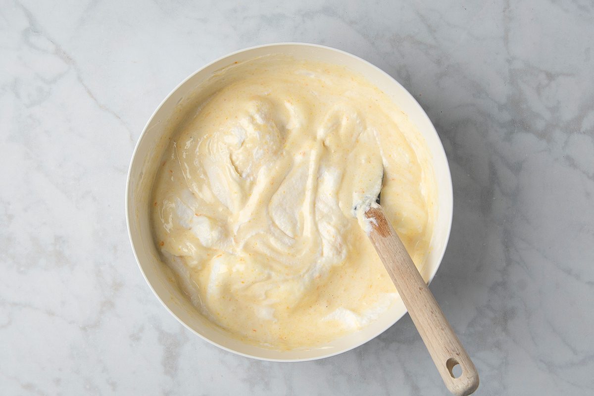 A white mixing bowl filled with pale yellow cake batter sits on a marble countertop. A wooden-handled spatula rests inside the bowl, partially covered in batter. The mixture has a smooth, creamy texture.