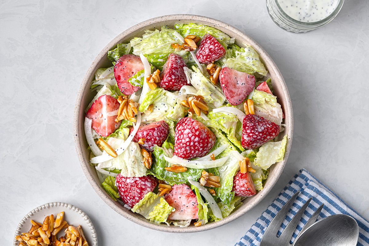 A bowl of salad with strawberries, romaine lettuce, and almonds on a light surface. A small dish of almonds and a blue-striped napkin with a fork and spoon are beside it. A small cup of dressing is in the background.